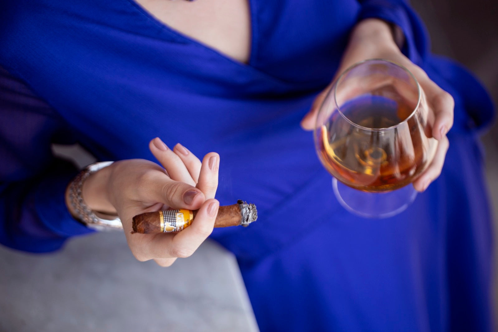 Woman in blue gown holding a short corona cigar and a glass of cognac in a moody lounge setting