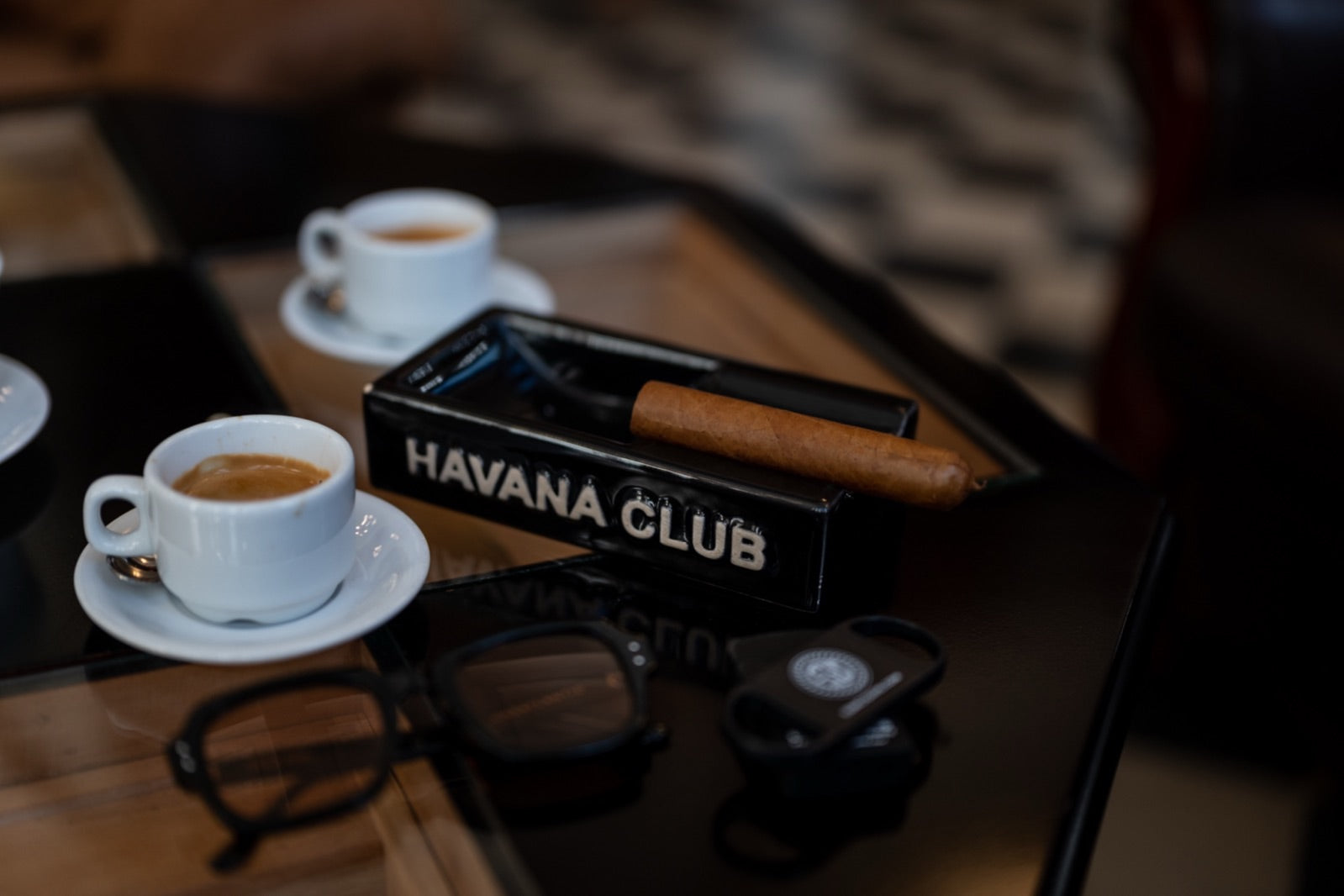 A single premium cigar resting in a Havana Club box beside two espressos, evoking the unhurried time it takes to smoke a cigar