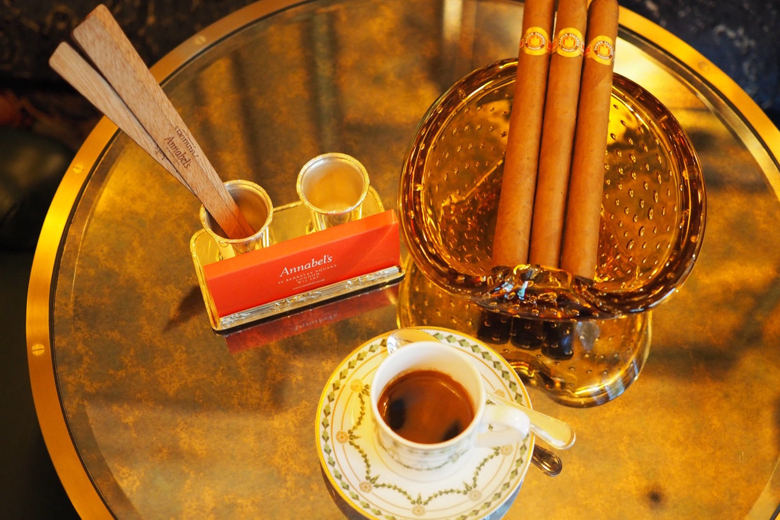 Three well-humidified cigars resting in an amber glass ashtray beside espresso on a gold table, illustrating how to store cigars