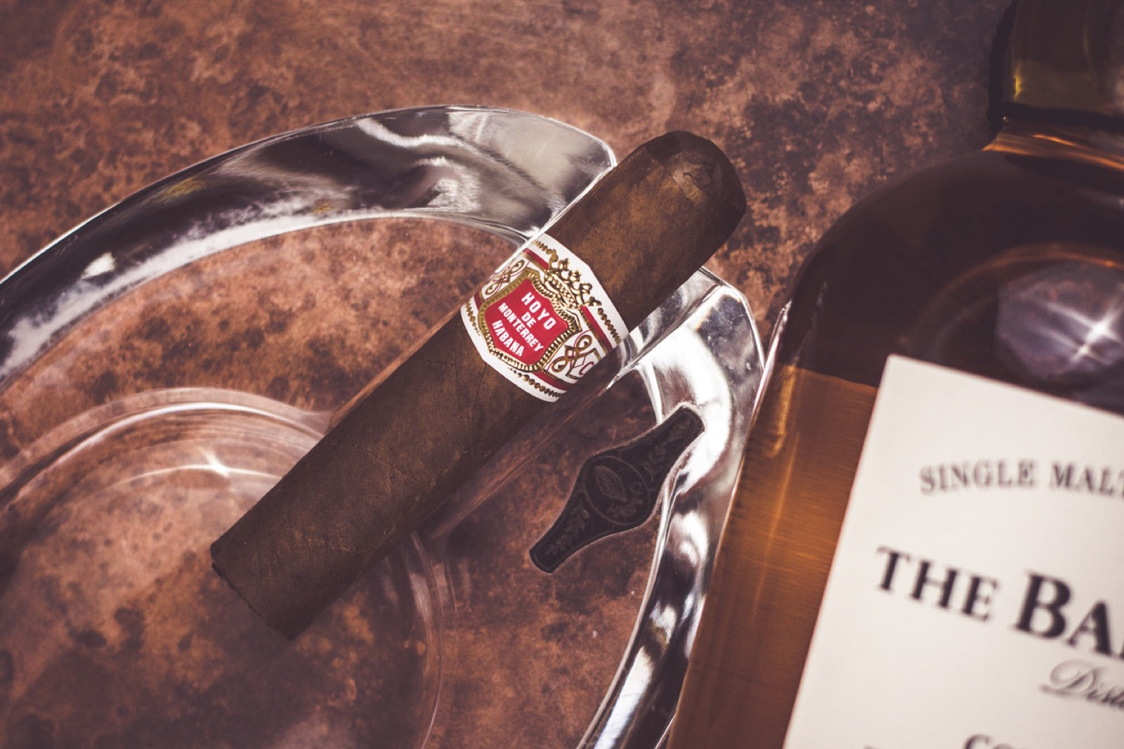 Hoyo de Monterrey Cuban cigar resting on a crystal ashtray beside a single malt whisky bottle in a moody lounge