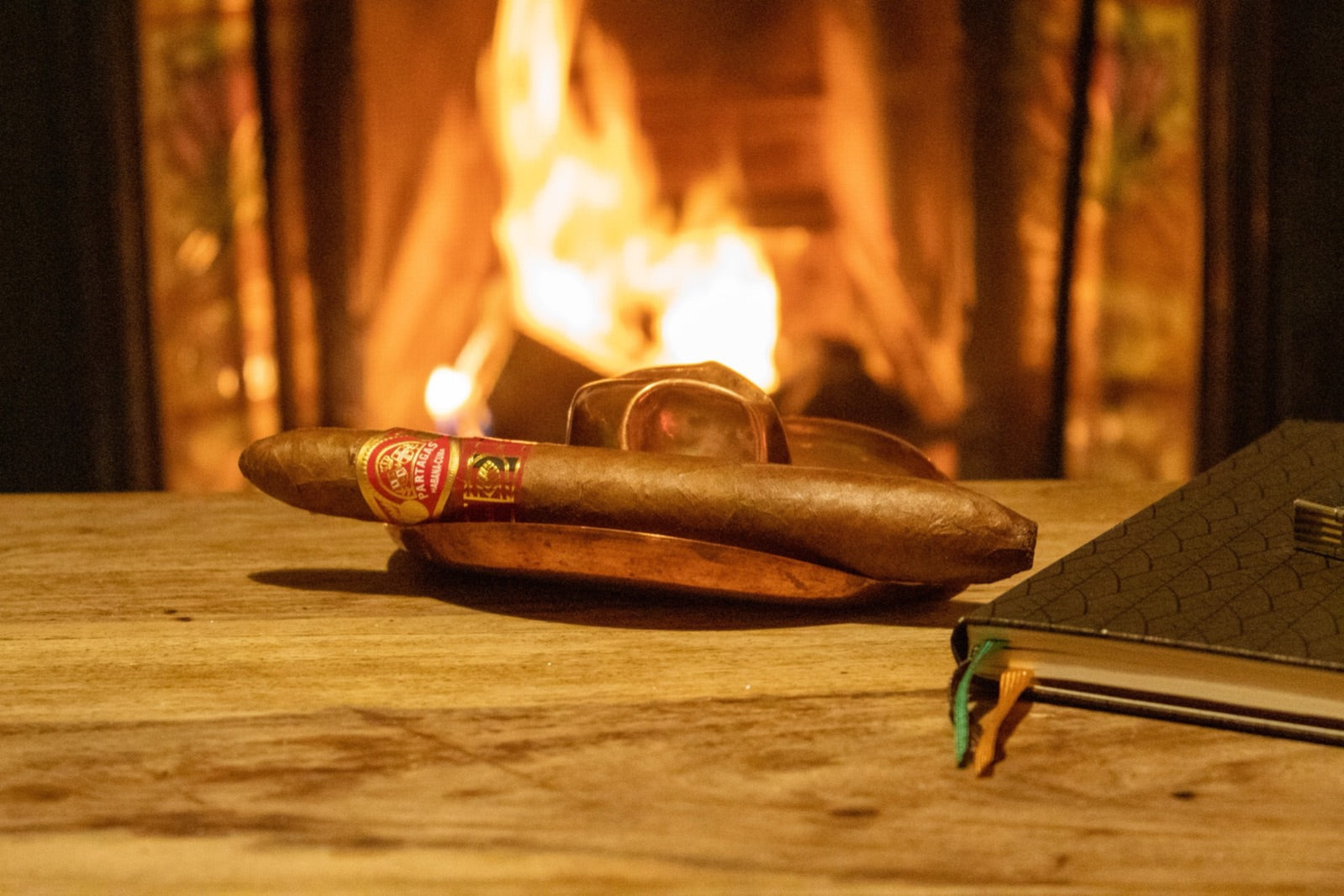 Partagas Salomones Cuban cigar resting on a copper ashtray before a roaring fireplace on a rustic wooden table
