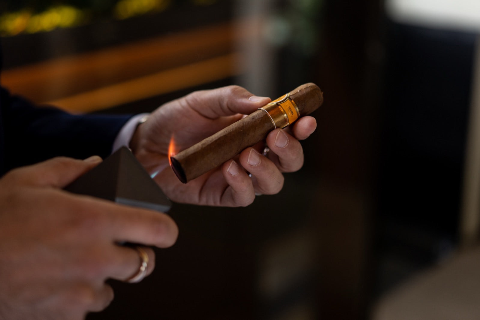 Man lighting a premium everyday cigar with a flame, gold band visible, in a warm luxury lounge setting