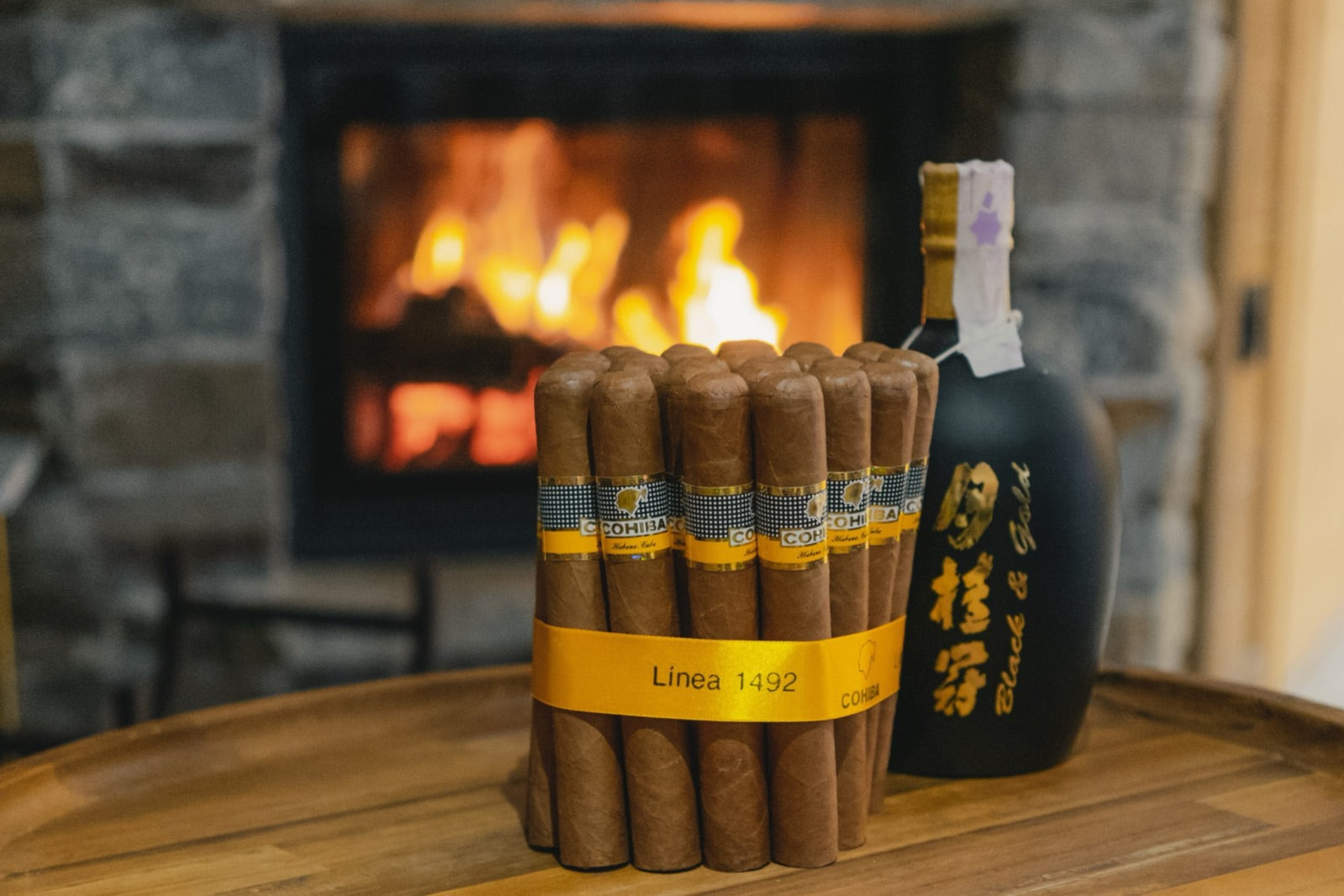 Bundle of Cohiba cigars beside a dark spirits bottle on a wooden table before a roaring stone fireplace, the classic cigars and cognac pairing