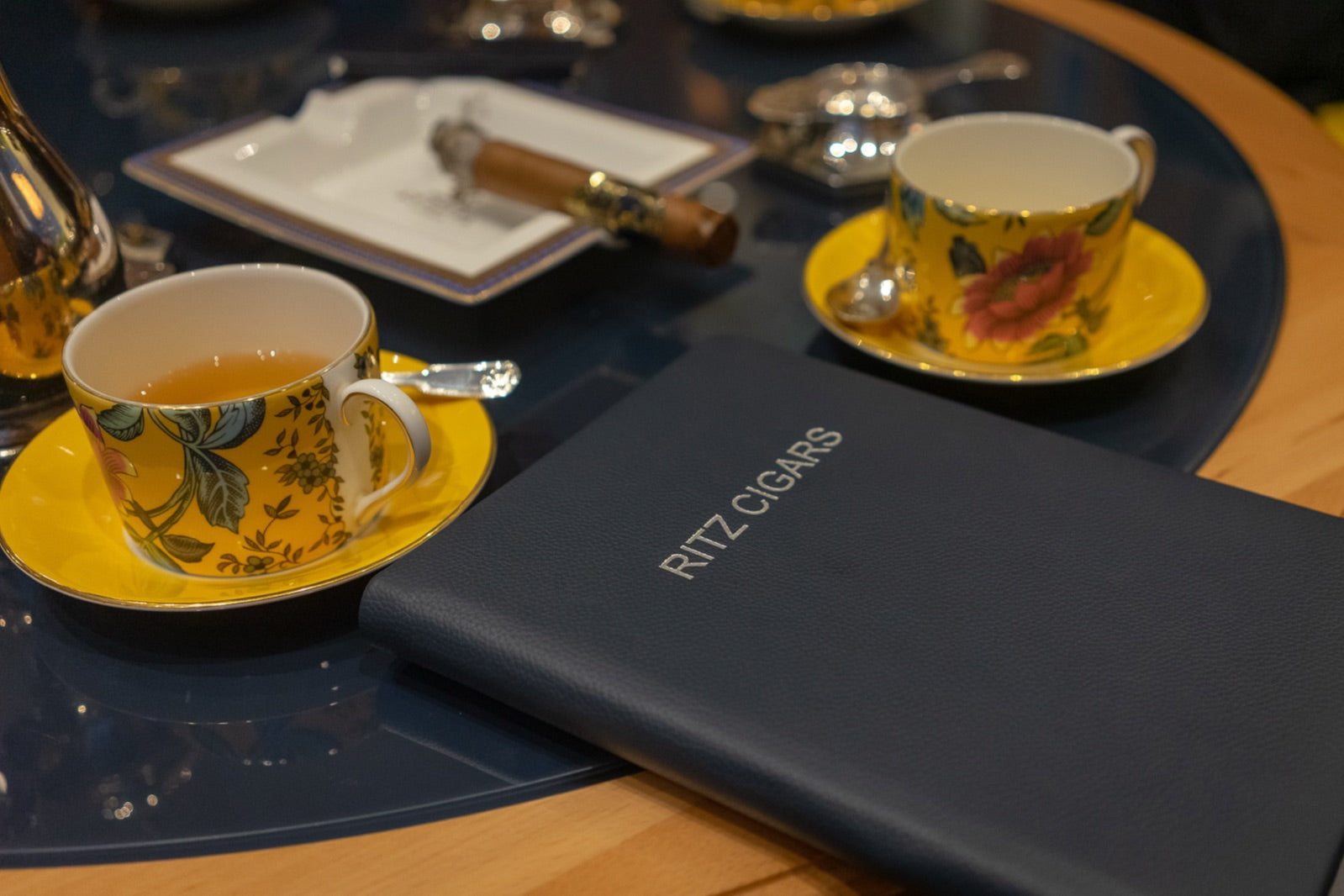 A cup of tea beside a fine cigar and a Ritz cigar menu on a polished table, pairing cigars and tea