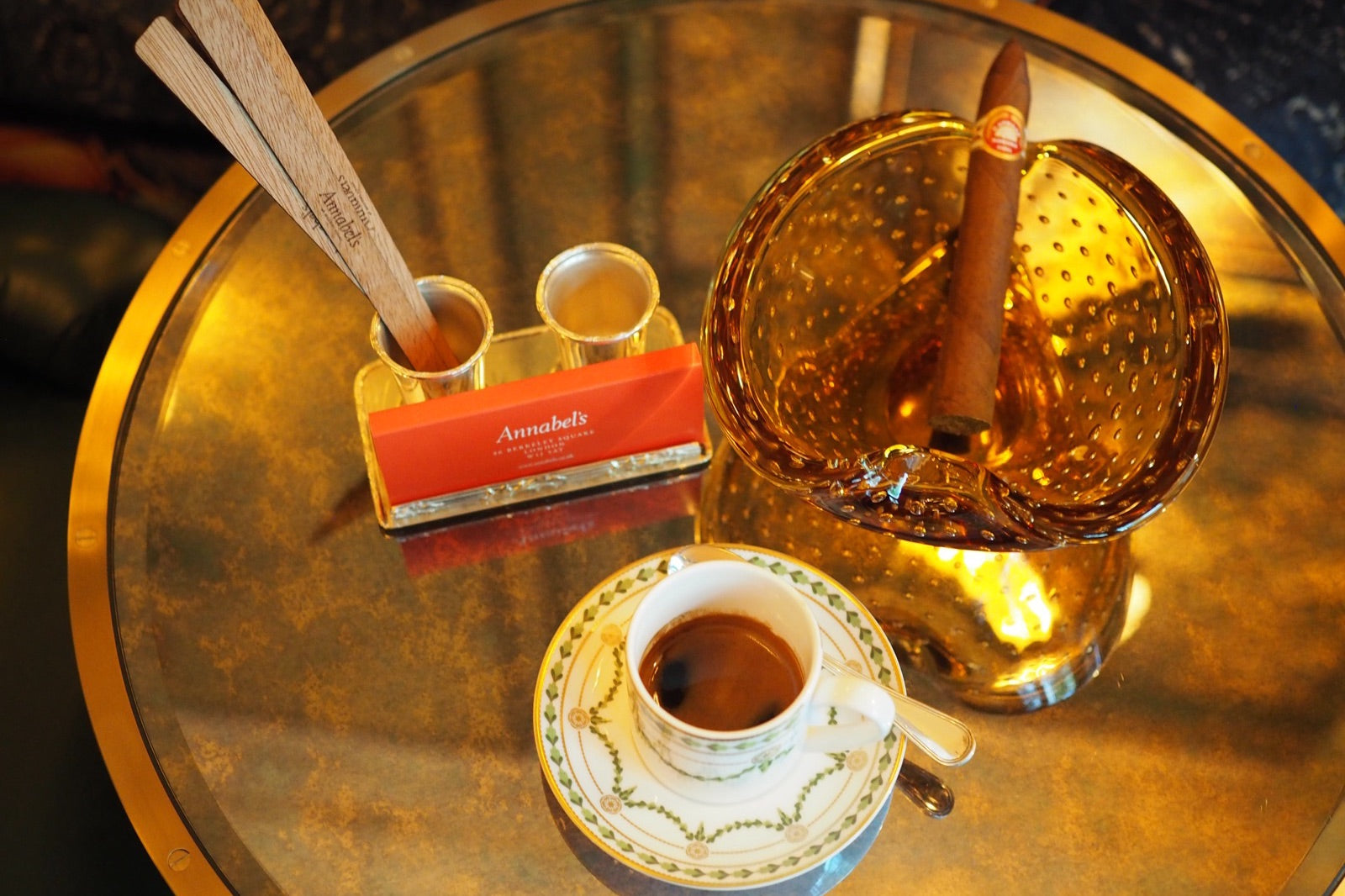 Lit Cuban cigar resting on a golden ashtray beside a drink on a gilded lounge table, evoking a cigars and whisky pairing