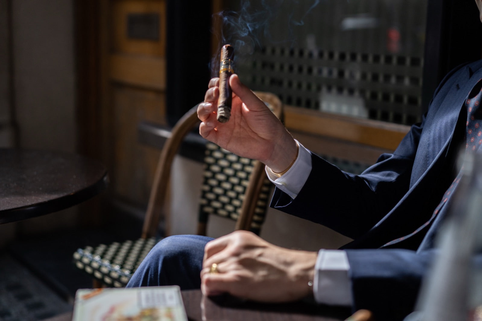 Hand of a man in a tailored suit holding a lit El Rey del Mundo cigar with smoke curling at a moody London cafe