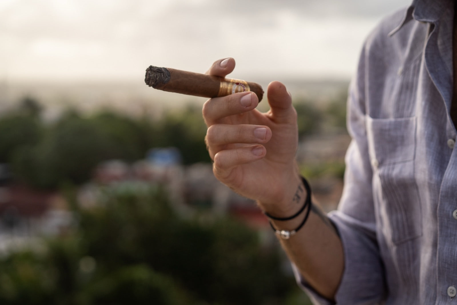 Hand holding a lit H. Upmann Cuban cigar against a soft green Havana balcony backdrop at dusk