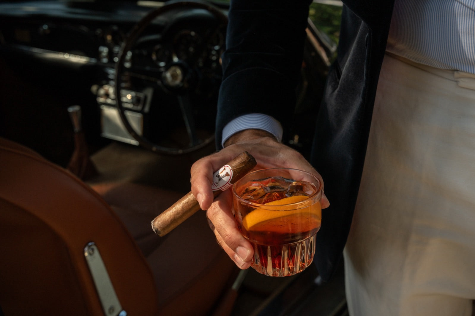 Hand holding a premium cigar and an amber cocktail beside a classic car, the perfect cigar gift for a client or colleague