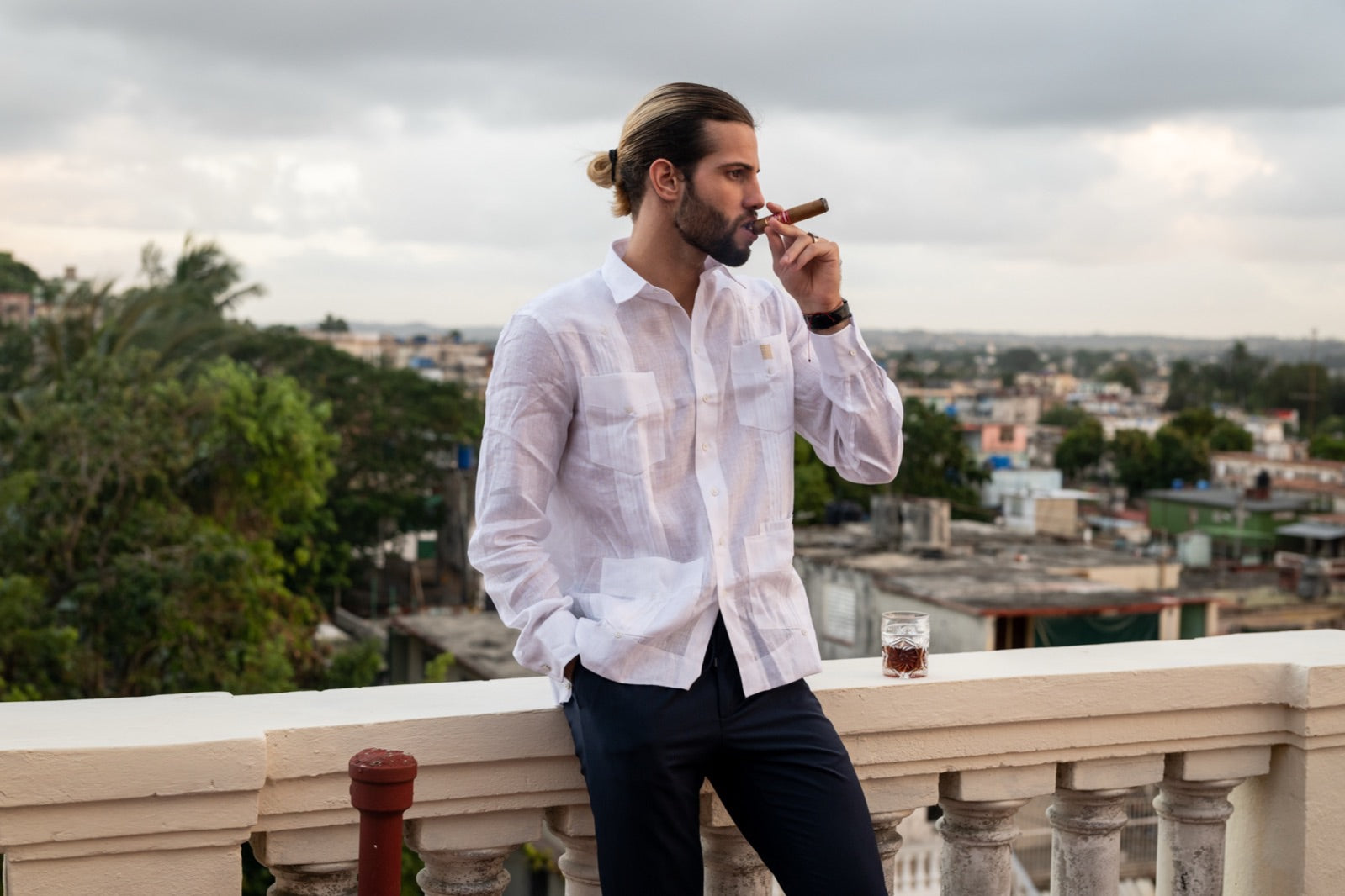 Man smoking a Cuban cigar with a glass of rum on a rooftop terrace overlooking the Havana skyline
